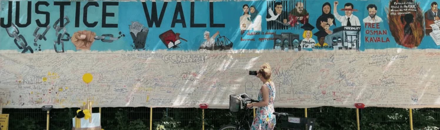 Une passante à vélo photographie le Mur de la justice : une longue fresque représentant des personnes emprisonnées, au-dessus d’une toile couverte de messages manuscrits.