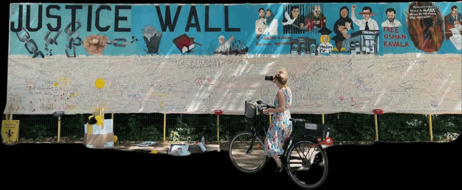 Une passante à vélo photographie le Mur de la justice : une longue fresque représentant des personnes emprisonnées, au-dessus d’une toile couverte de messages manuscrits.